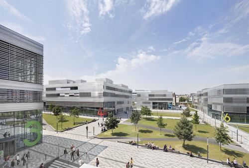 Moderner Hochschulcampus mit mehreren silbernen Gebäuden, die um einen begrünten Innenhof mit Wegen, Bäumen und Sitzgelegenheiten angeordnet sind. Studierende bewegen sich über den Platz oder sitzen in kleinen Gruppen im Freien.